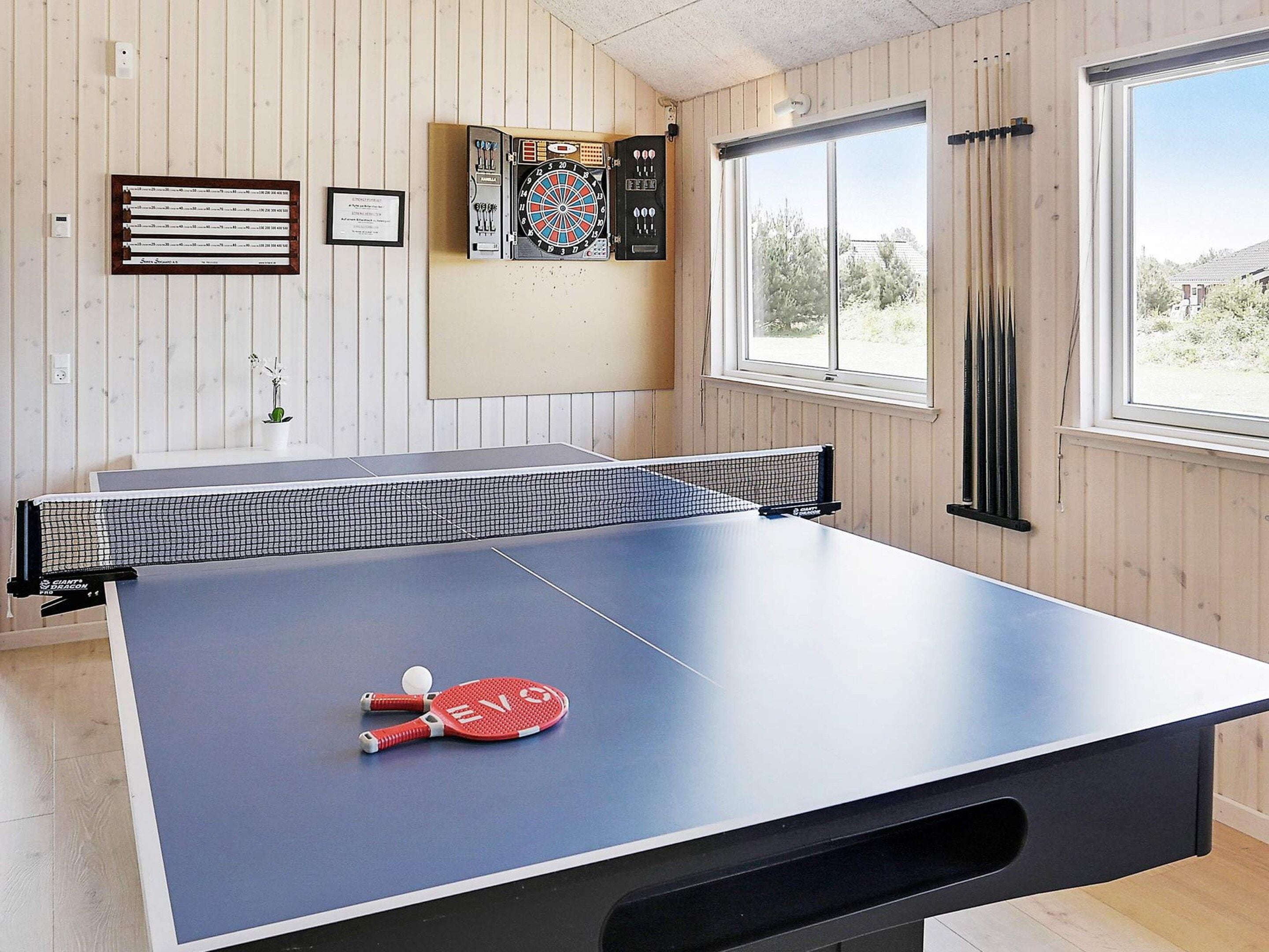 A ping pong table with a net, a red paddle, and a ball. There is a dartboard and a framed certificate on the wall.