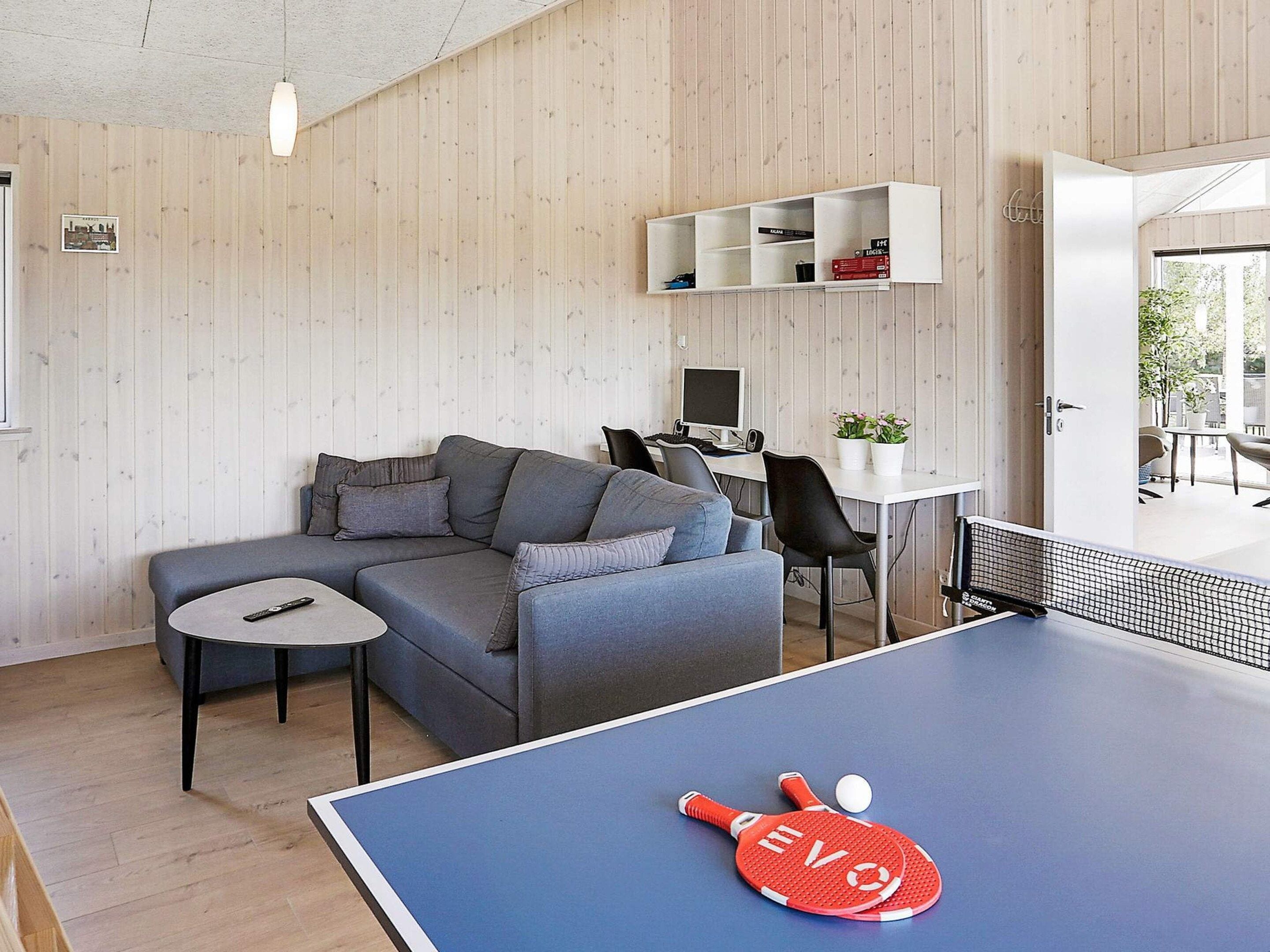 A modern living room with a blue ping pong table, grey sofa, and a white desk with a computer.