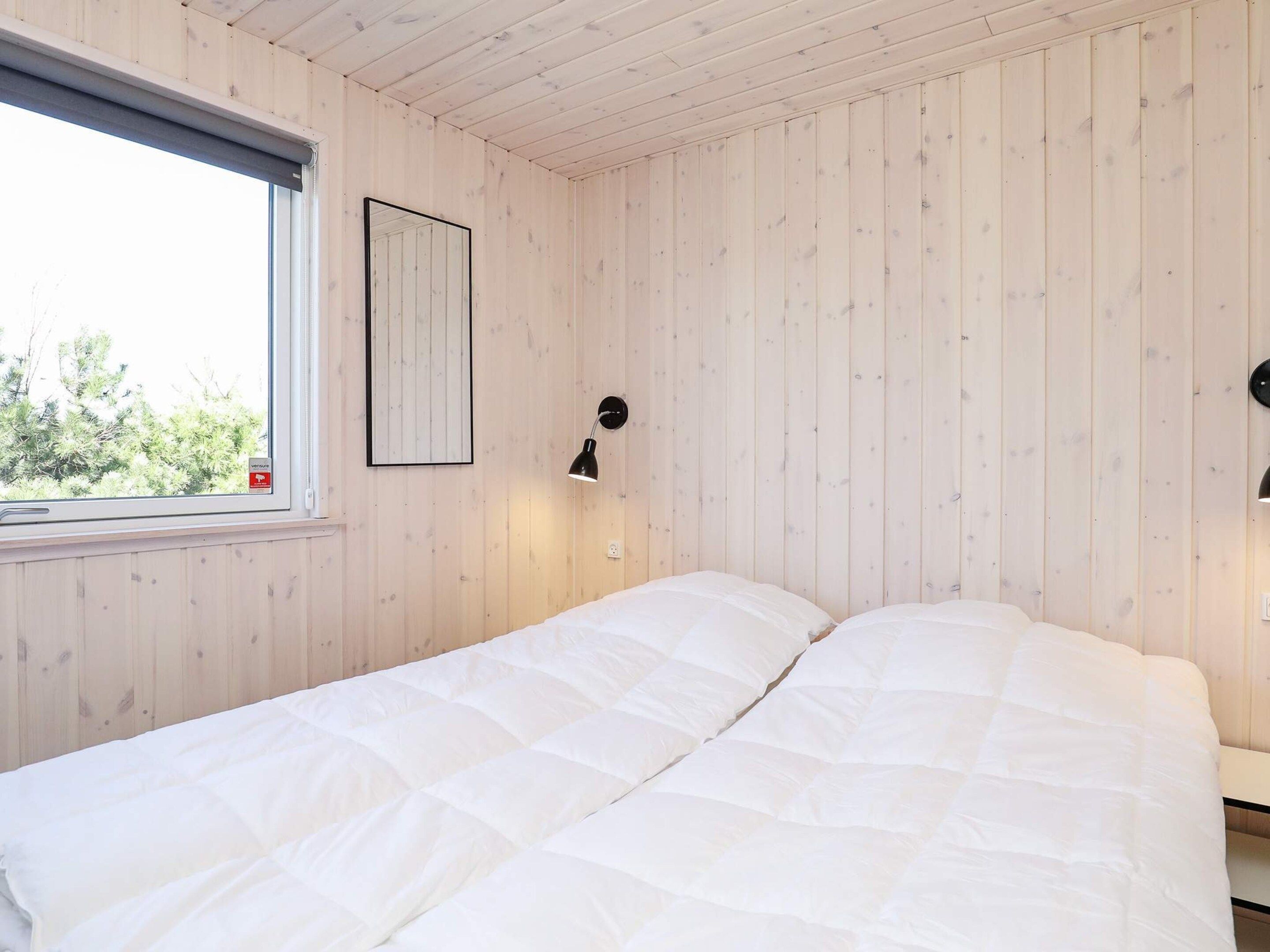 A room with wooden panel walls, a bed with white bedding, a framed picture on the wall, and a window with a view of trees.