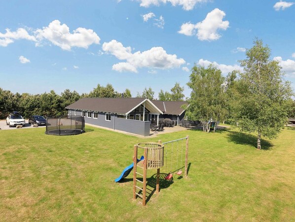 A playground with a slide, swings, and a trampoline in a grassy area with a building in the background. - Luxury Holiday Home in Falster near Sea (Vaeggerlose)