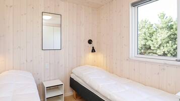 A room with two beds, a small white nightstand, a wall-mounted mirror, a window with a view of trees, and a wall-mounted lamp.