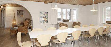 A spacious dining area with a long table and multiple chairs, wooden ceiling, and large windows.
