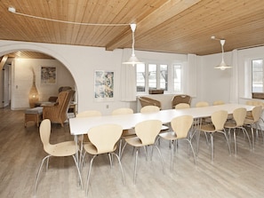 A spacious dining area with a long table and multiple chairs, wooden ceiling, and large windows.