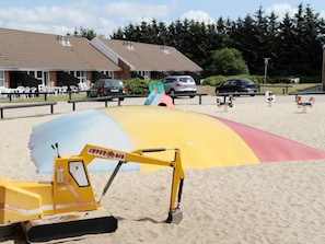 A yellow excavator on a sandy surface with a colorful slide structure in the background.