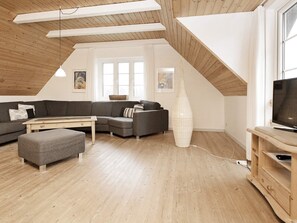A loft-style living room with a wooden ceiling, a large window, a gray sectional sofa, a white vase, and a television on a wooden stand.