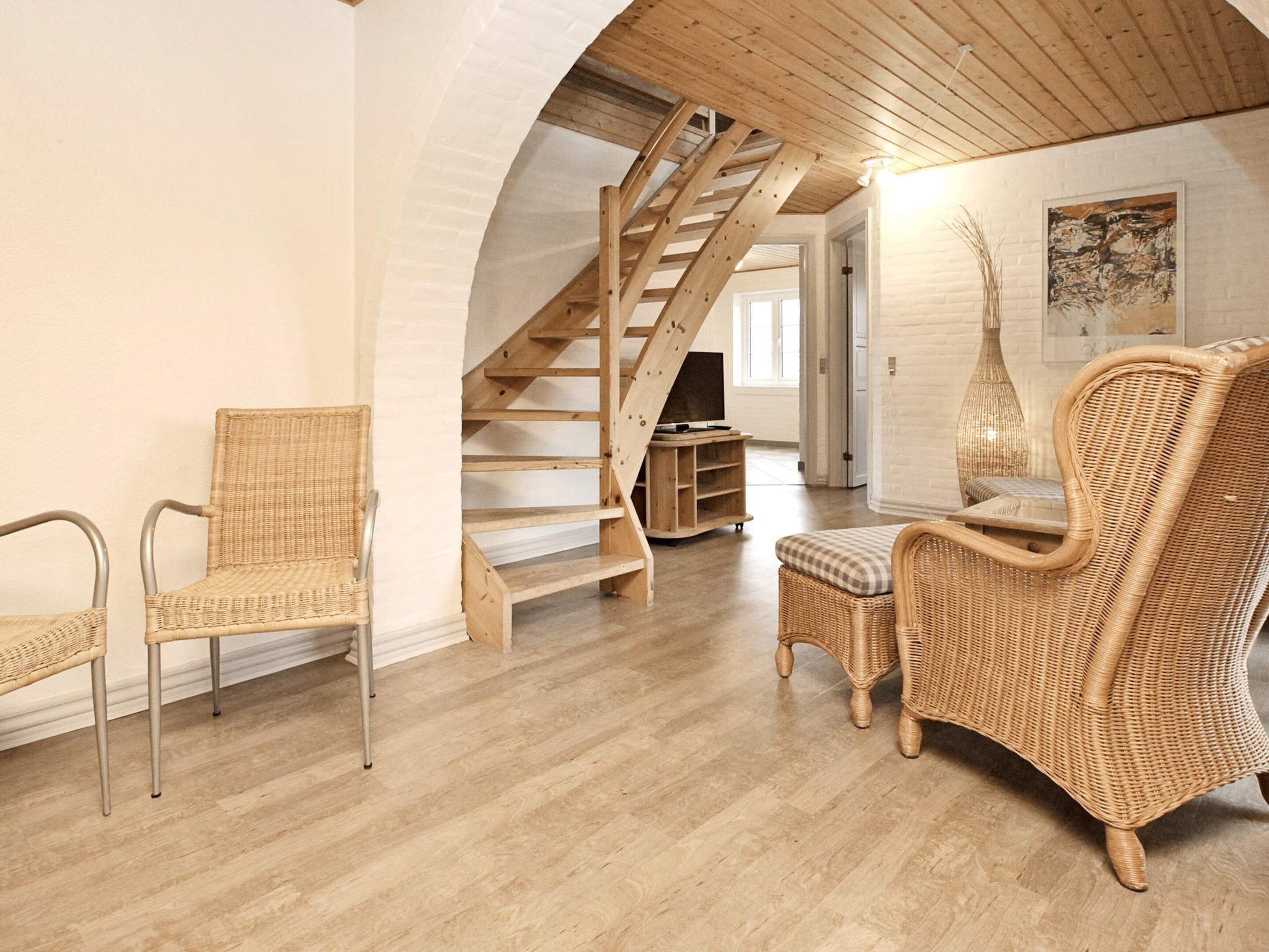 Una habitación amplia con escalera de madera, muebles de mimbre y pared de ladrillo blanco.