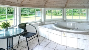 A covered outdoor area with a hot tub, a table with a chair, and large windows offering a view of greenery. - 12 Person Holiday Home in Vaeggerlose (Vaeggerlose)