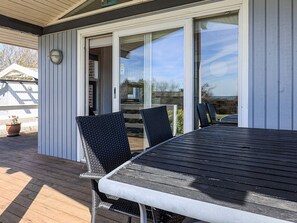 Une terrasse extérieure couverte, avec une table et des chaises, une porte-fenêtre coulissante donnant sur une terrasse, et un pot de fleurs.