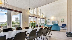 Ein geräumiger Essbereich mit einem langen Tisch und modernen Stühlen, eine blaue Couch und ein Fernseher in einem Wohnbereich mit Holzwänden und großen Fenstern.