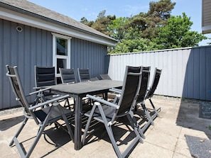 An outdoor patio with a metal table and chairs, a window, and a grey building in the background.