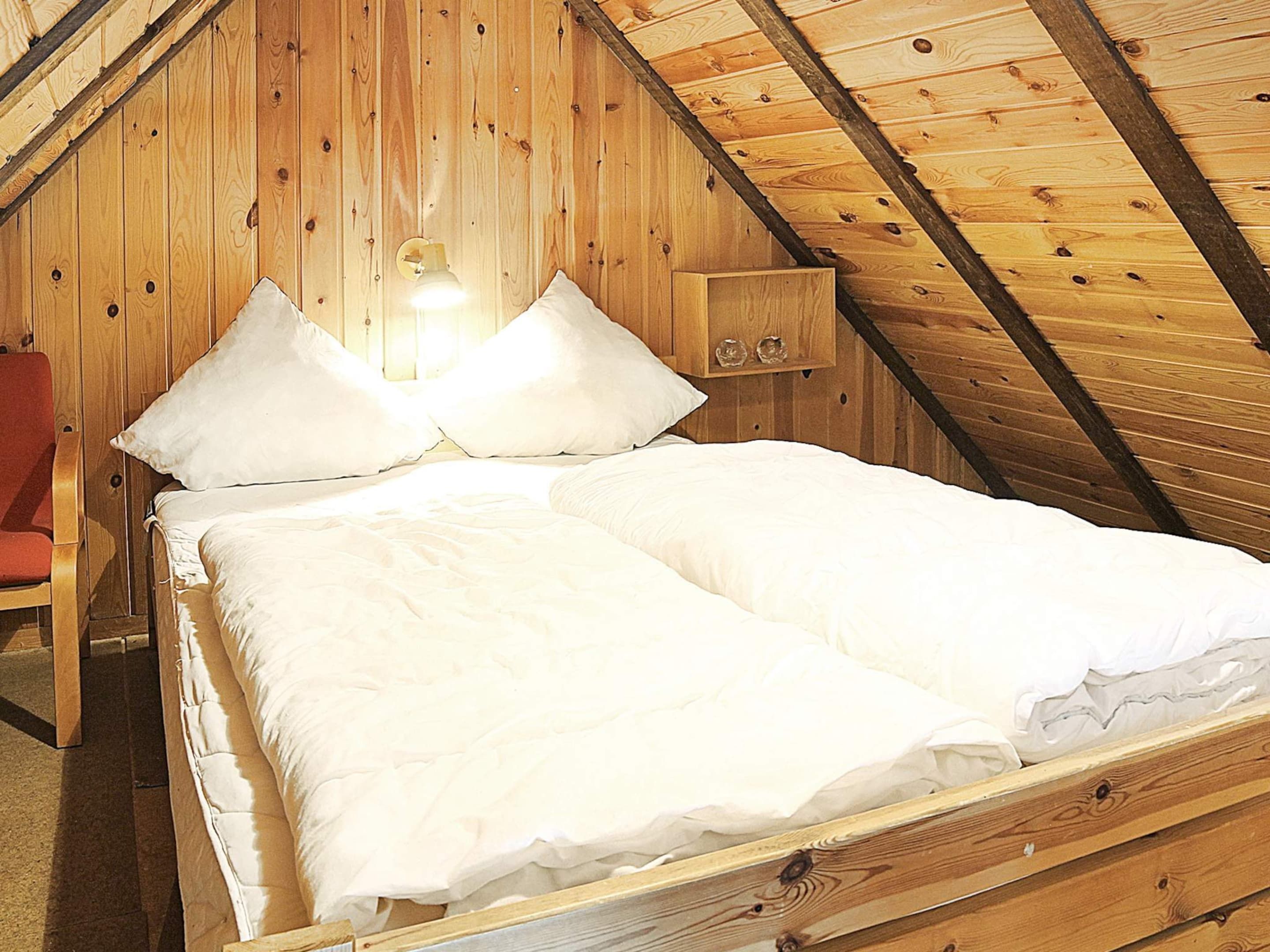 A single bed in a wooden room with a sloped ceiling, a nightstand with a lamp, and a red chair.