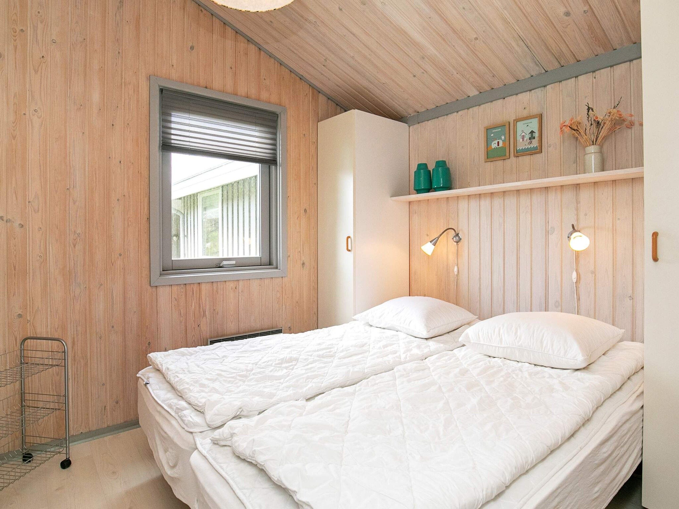 A bedroom with a wooden ceiling, a bed with white bedding, a window with blinds, and a shelf with green objects.