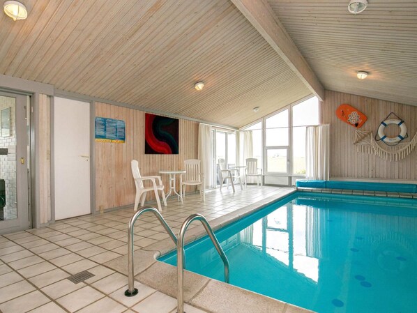 An indoor swimming pool with tiled flooring, a pool ladder, and white plastic chairs and tables.