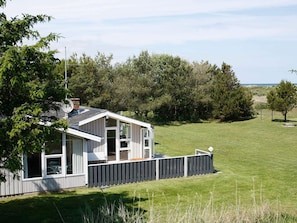 A single-story house with a fenced deck, surrounded by a grassy lawn and dense trees.