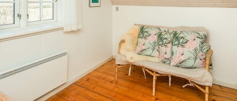 A room with a sloped ceiling, a window with white curtains, a radiator, a wicker sofa with a floral cushion, and a framed picture on the wall.