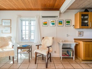 Una sala de estar acogedora con techo de madera, una silla blanca con un cojín mullido, una mesa de madera con libros, una ventana con cortinas blancas y una pared decorada con obras enmarcadas.