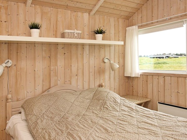 Una habitación con paredes de madera, una cama con un edredón estampado, una ventana que ofrece vistas al exterior y un estante con plantas y una cesta.
