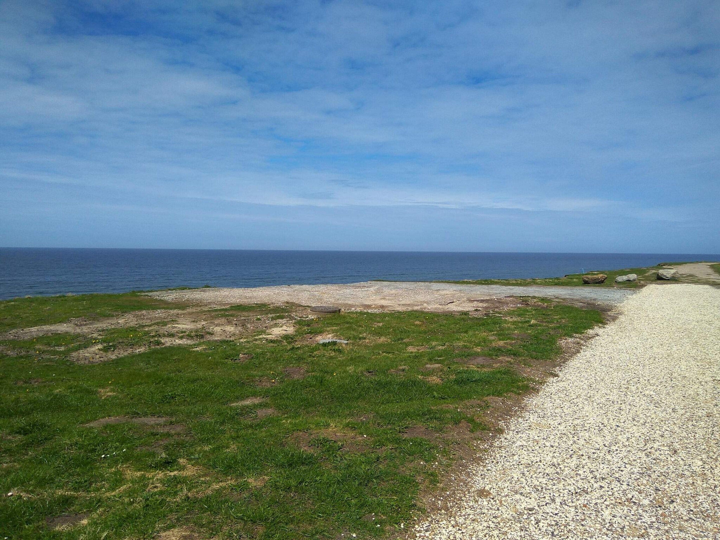 A coastal landscape with a gravel path leading to the sea, grassy areas, and scattered rocks.