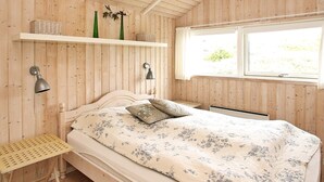 A room with a wooden ceiling, a bed with floral bedding, two wall-mounted lamps, a window with a view, and shelves with decorative items. - Quaint Holiday Home in LÃ¸kken near Beach (Lokken)