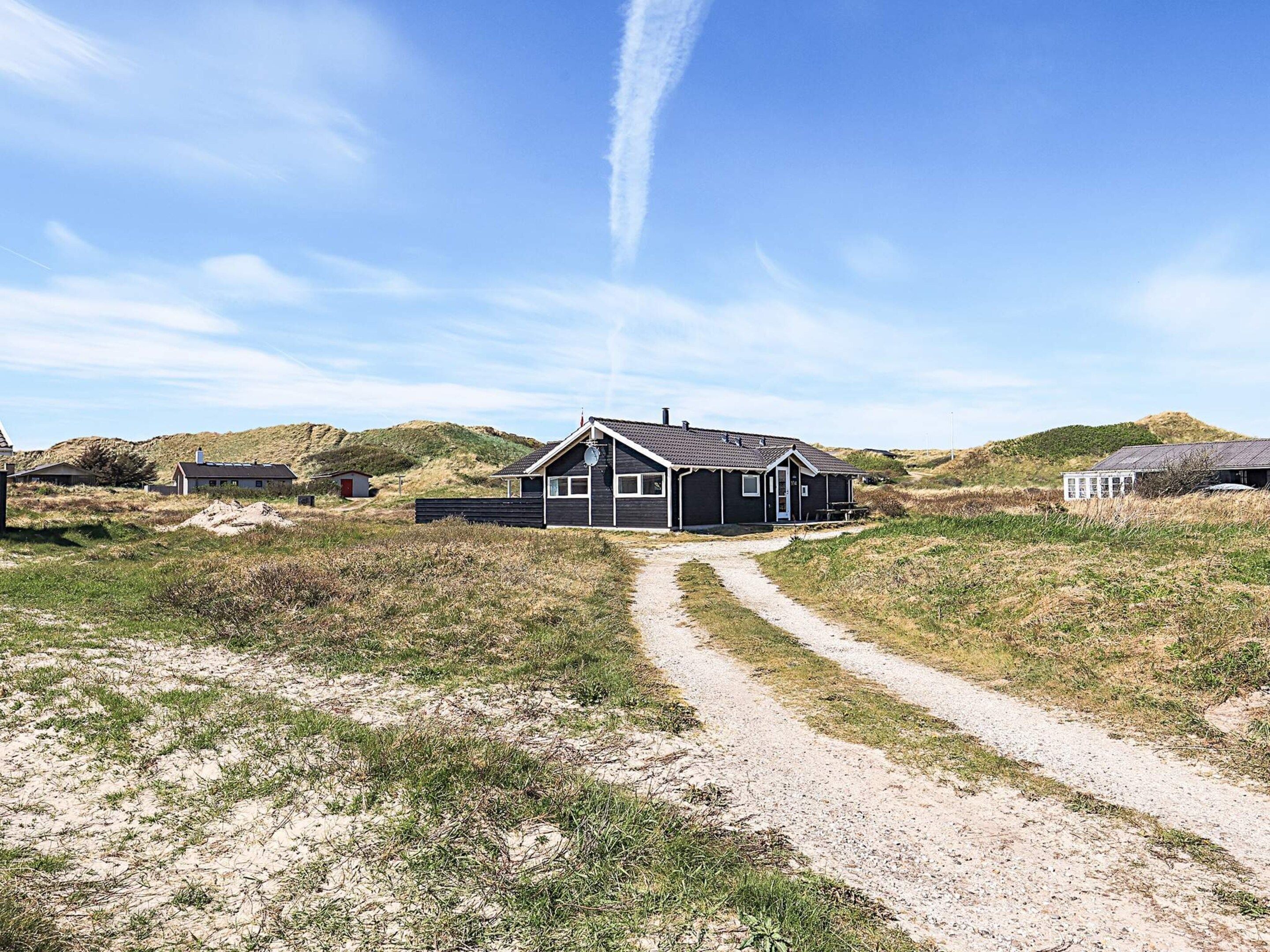 Ein einstöckiges Gebäude mit Kiesweg, umgeben von grasbewachsenen Dünen und blauem Himmel.