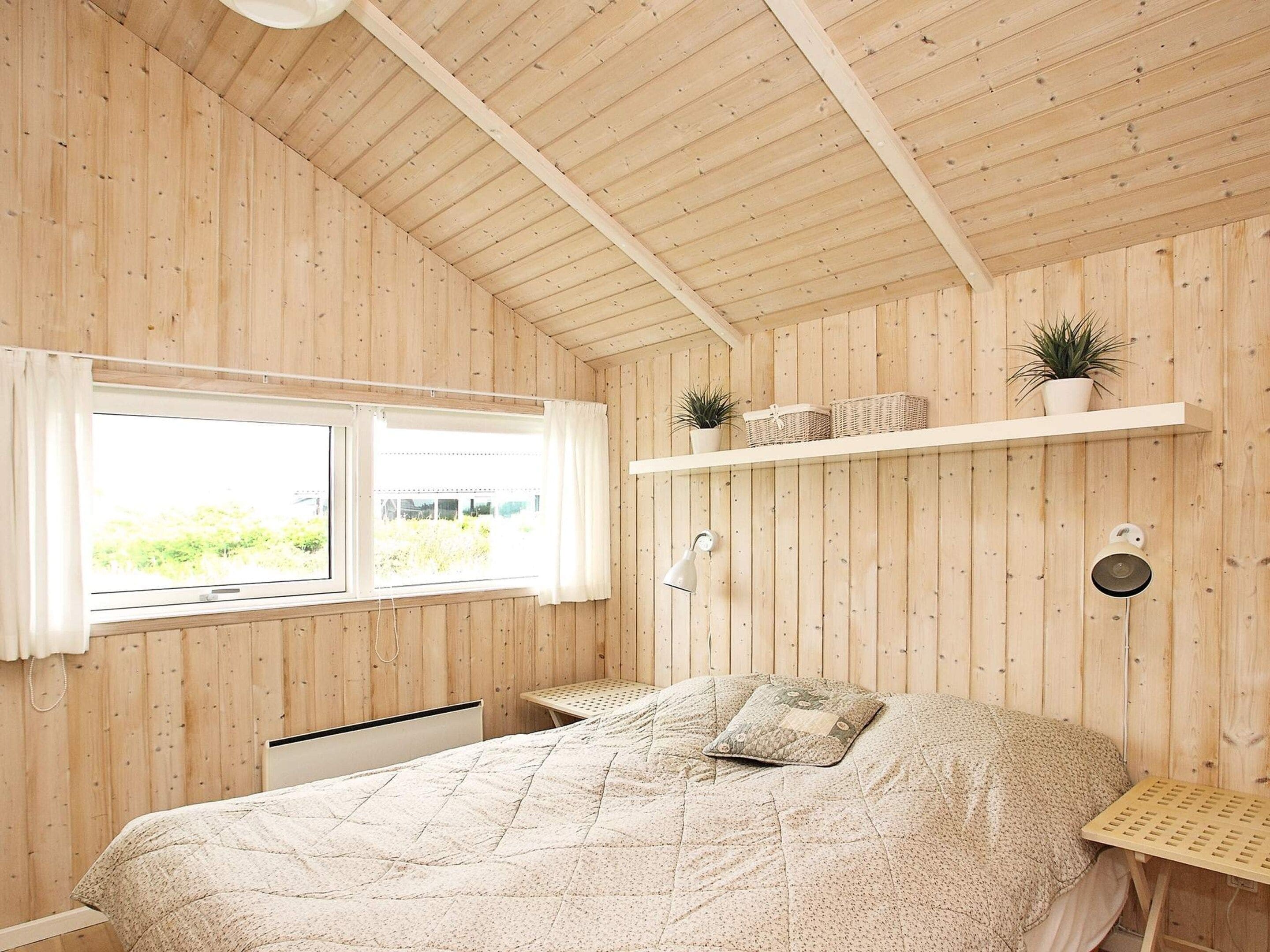 A bedroom with a bed, a window, a shelf with plants, and a wall-mounted lamp.