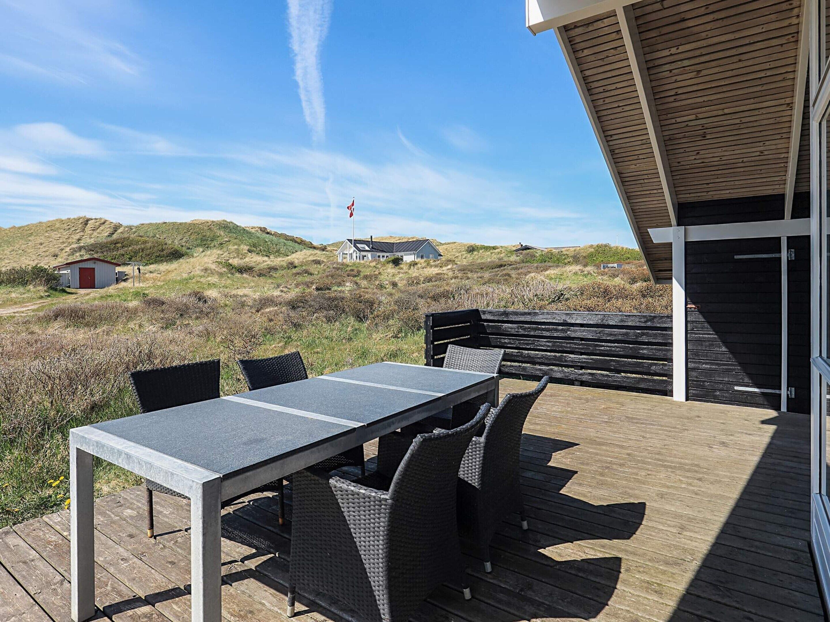 Una terraza exterior con mesa y sillas, con vista a una playa con dunas y un asta de bandera.