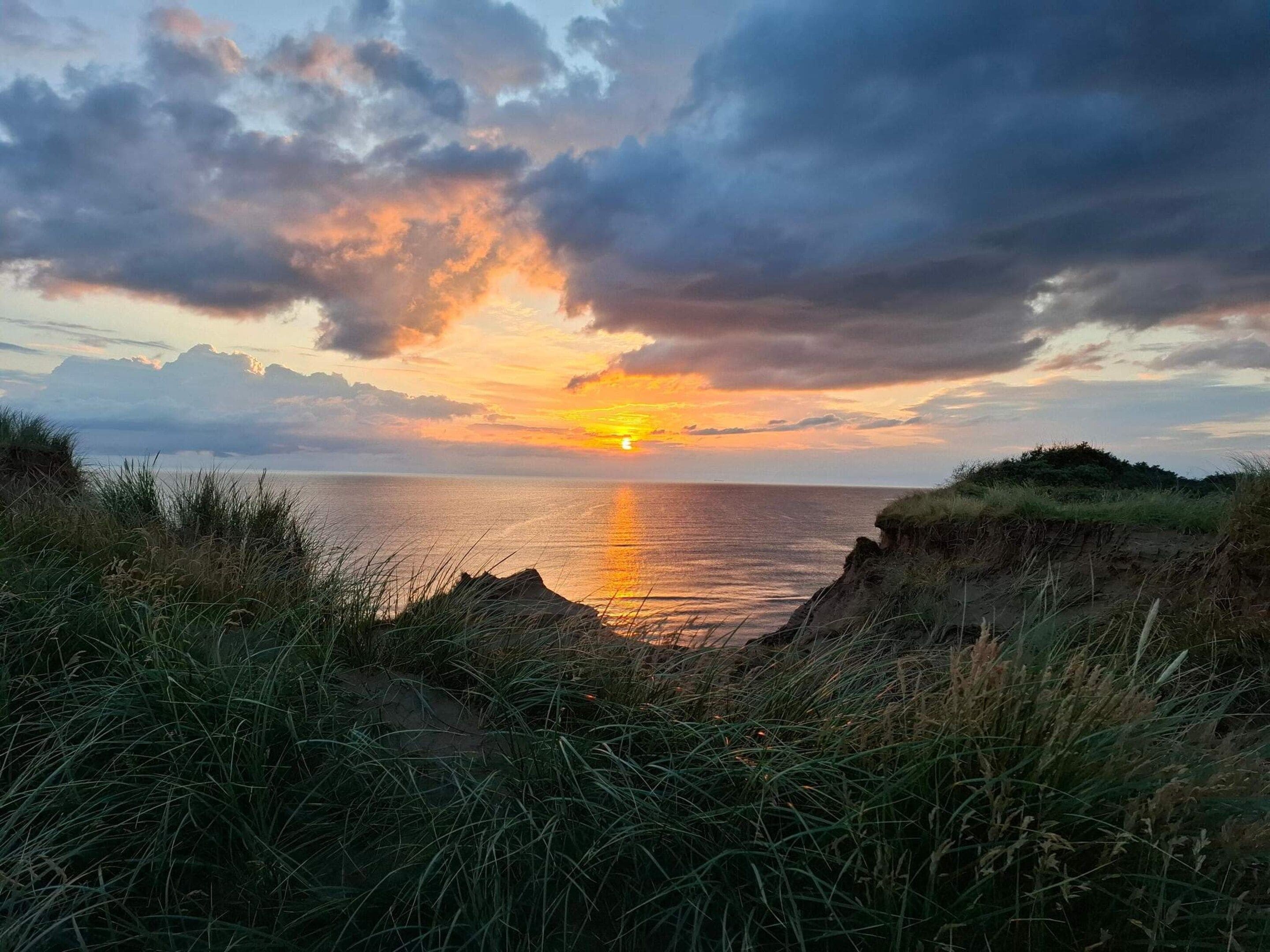 A coastal sunset with a clear view of the sea and a grassy foreground.