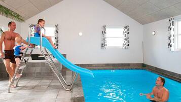 En indendørs swimmingpool med rutsjebane, flisegulv og vinduer.