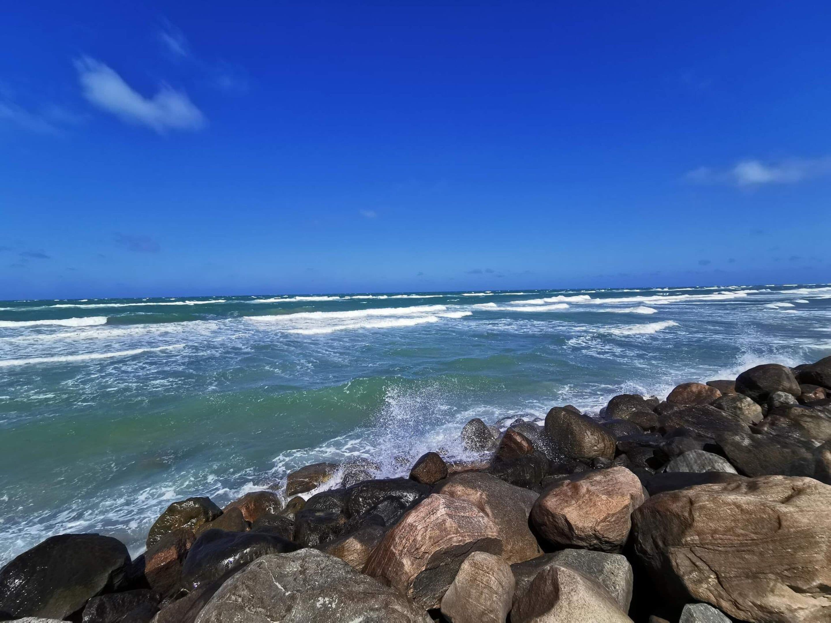Un rivage rocheux où les vagues s’écrasent contre les rochers, sous un ciel bleu dégagé.