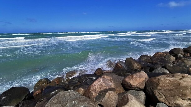Una costa rocosa con olas rompiendo contra las rocas bajo un cielo azul despejado.