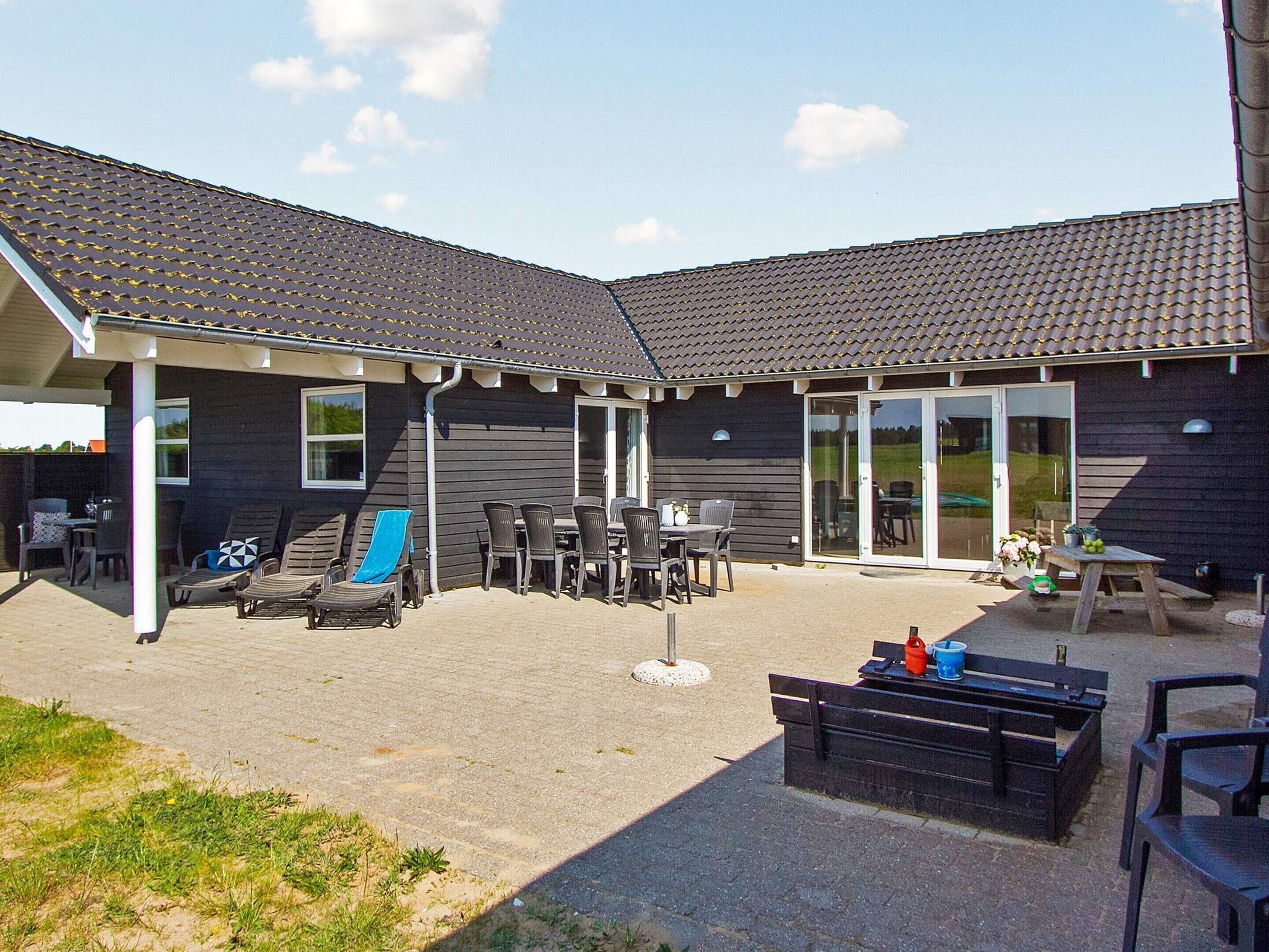 En overdækket patio med udendørs møbler og et bord, en bygning med et tegltag, og en dør der fører til en overdækket indgang.