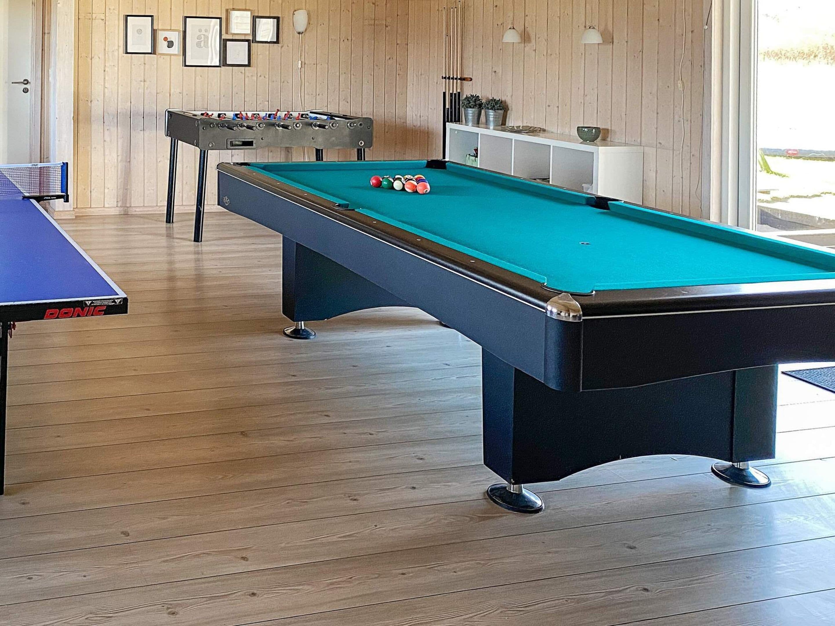 Una mesa de billar con superficie de fieltro azul, una mesa de fútbol de mesa y una mesa de billar en una habitación con suelo de madera y pared blanca decorada con cuadros.
