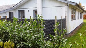 Un bâtiment blanc avec un toit en pente, une cheminée et une zone délimitée par une clôture, où poussent des arbustes verts.
