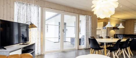 A modern living room with a flat-screen TV, leather sofa, dining area with chairs, and a large window with curtains.