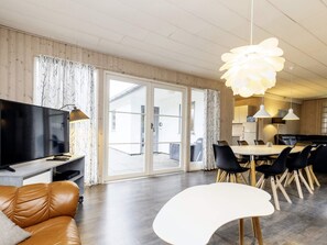 A modern living room with a flat-screen TV, leather sofa, dining area with chairs, and a large window with curtains. - Enticing Holiday Home in Falster near Sea (Vaeggerlose)