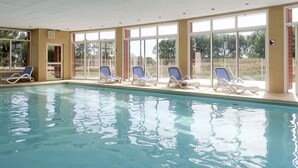 Ein Innenpool mit klarem, blauen Wasser, umgeben von weißen Liegestühlen und großen Fenstern.