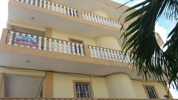 Um edifício de vários andares com varandas, um letreiro com 'RES. ROBMAR' e uma palmeira em primeiro plano.