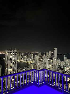 Une vue nocturne d’une ville, avec ses gratte-ciel, son front de mer et le garde-corps d’un balcon illuminé de lumière bleue.