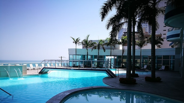 Piscina com espreguiçadeiras e palmeiras, cercada por edifícios modernos.