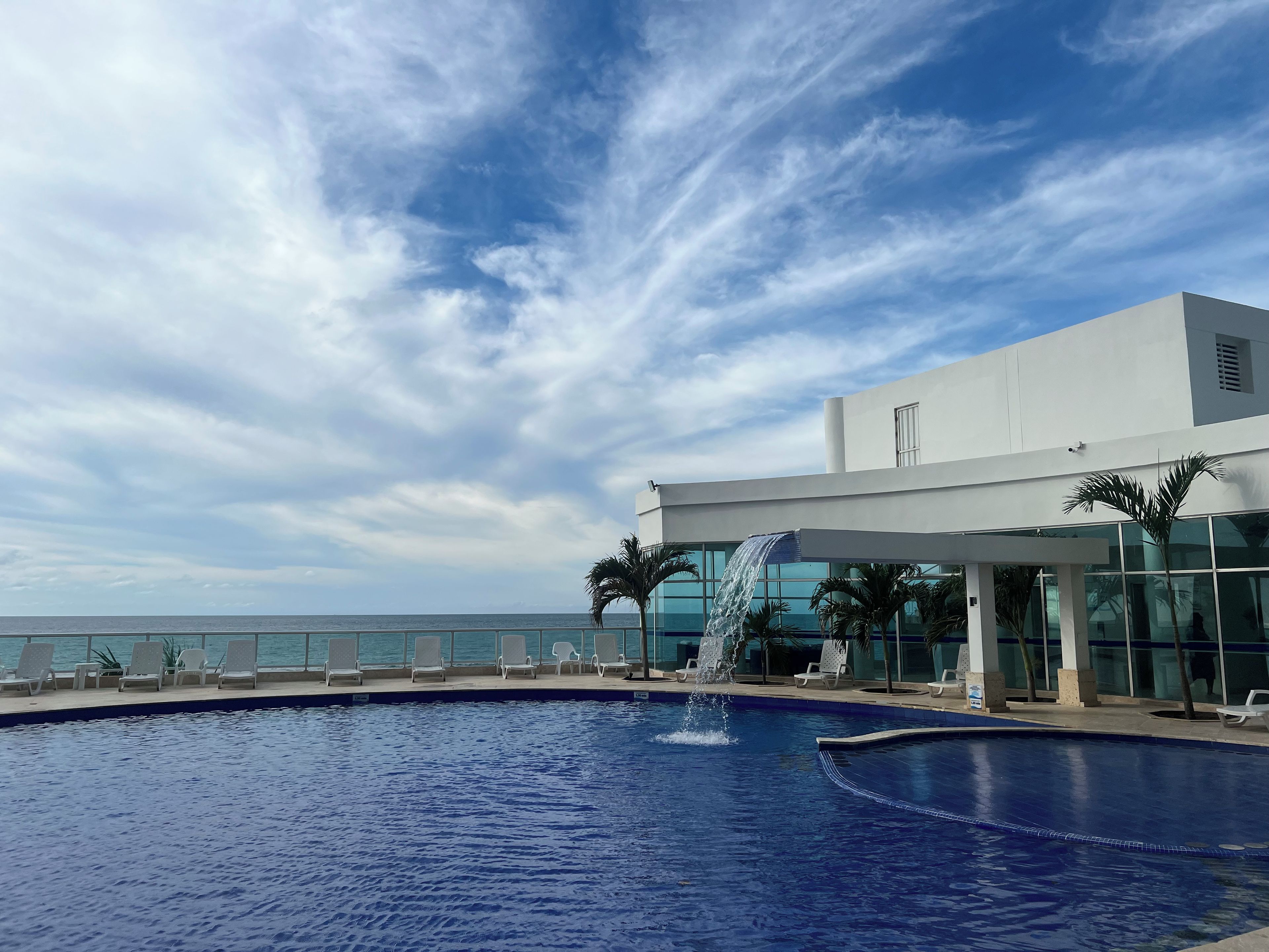 A swimming pool with a water feature, surrounded by lounge chairs and palm trees, with a modern building in the background.