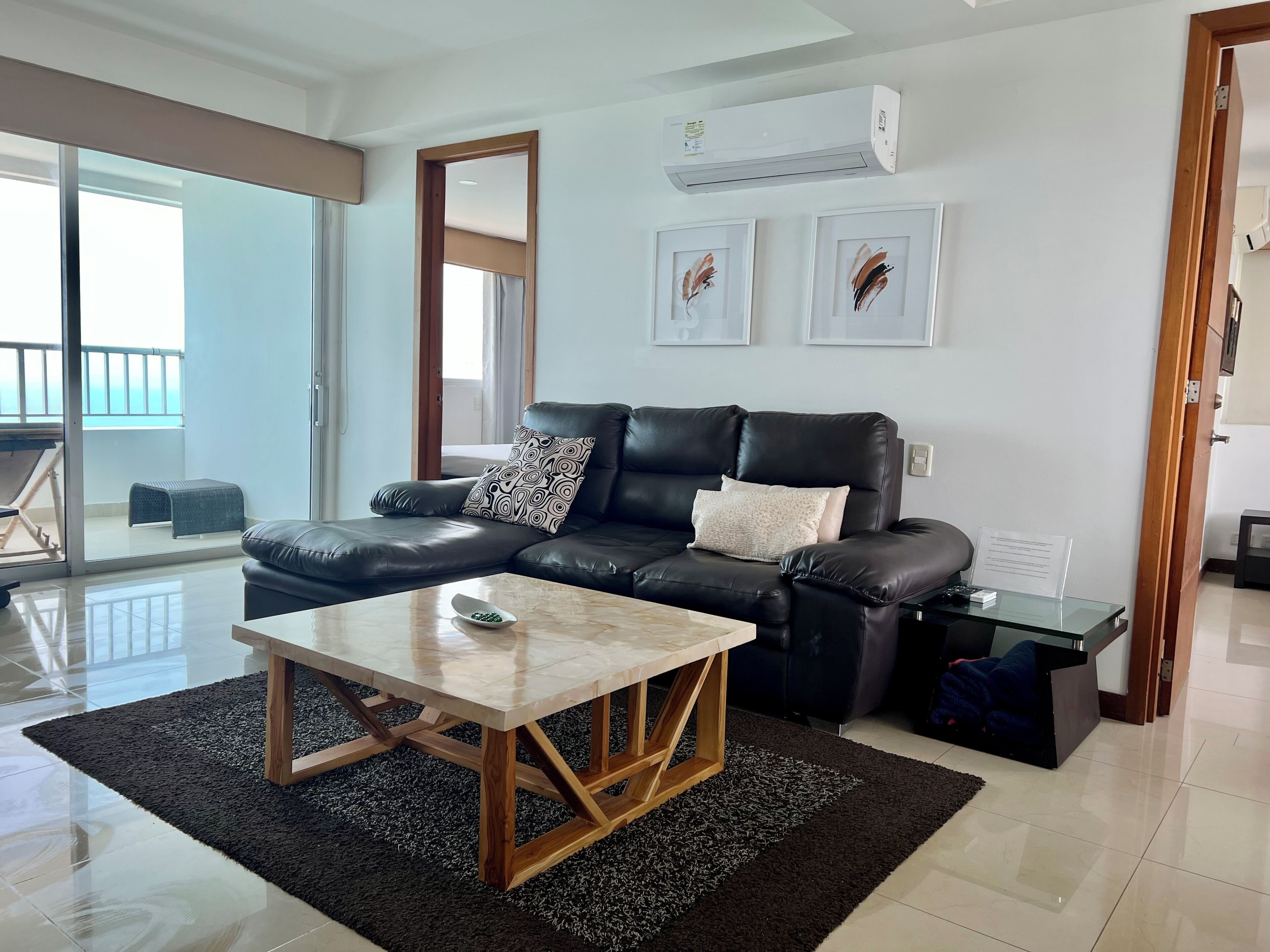 Uma sala de estar moderna com um sofá de couro preto, uma mesa de centro de madeira e uma mesa de jantar de vidro. Há uma janela grande com vista para o exterior, um aparelho de ar condicionado na parede e obras de arte emolduradas acima do sofá.