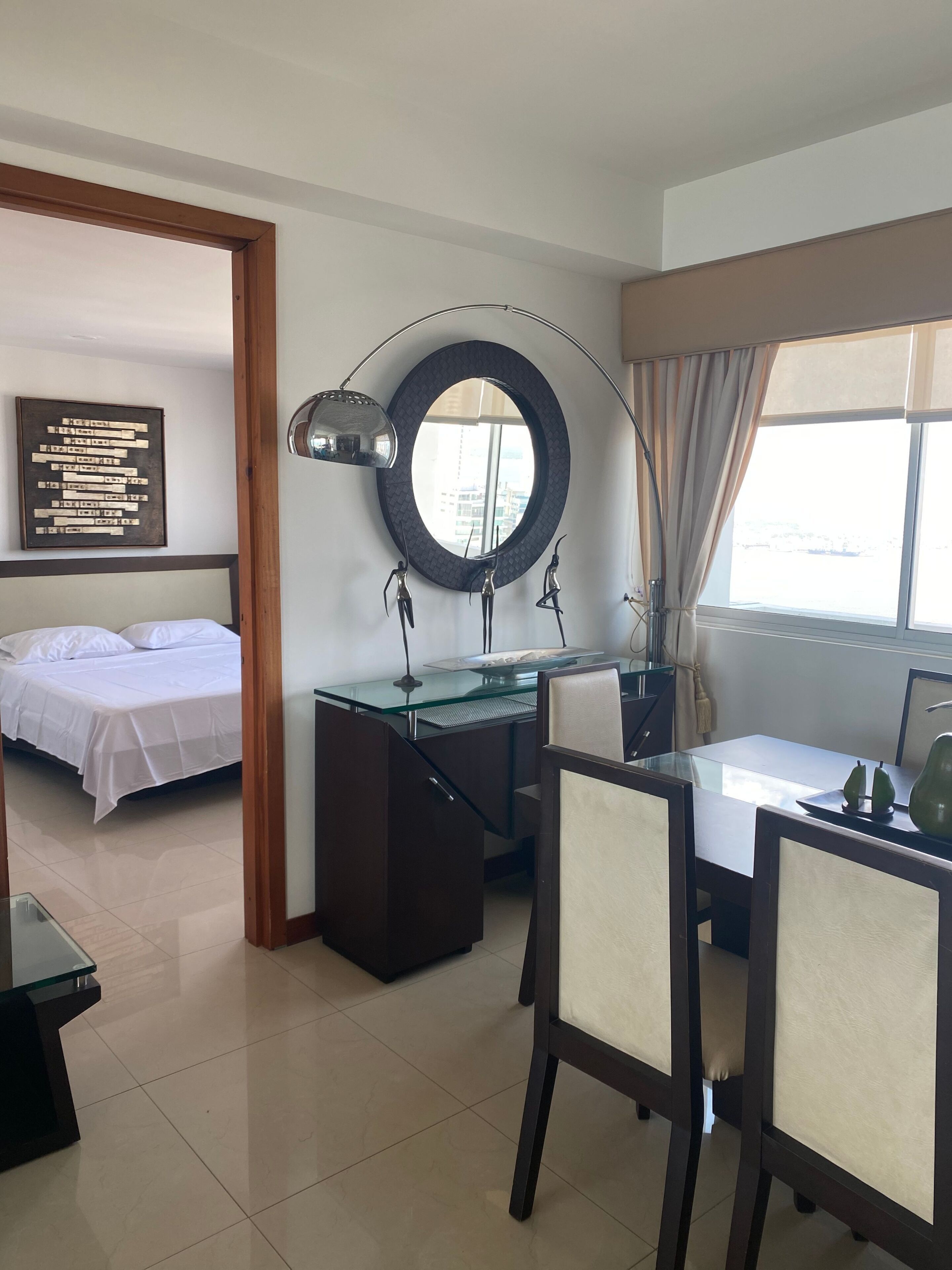 A hotel room with a bed, a desk with a chair, a round mirror, and a window with curtains.
