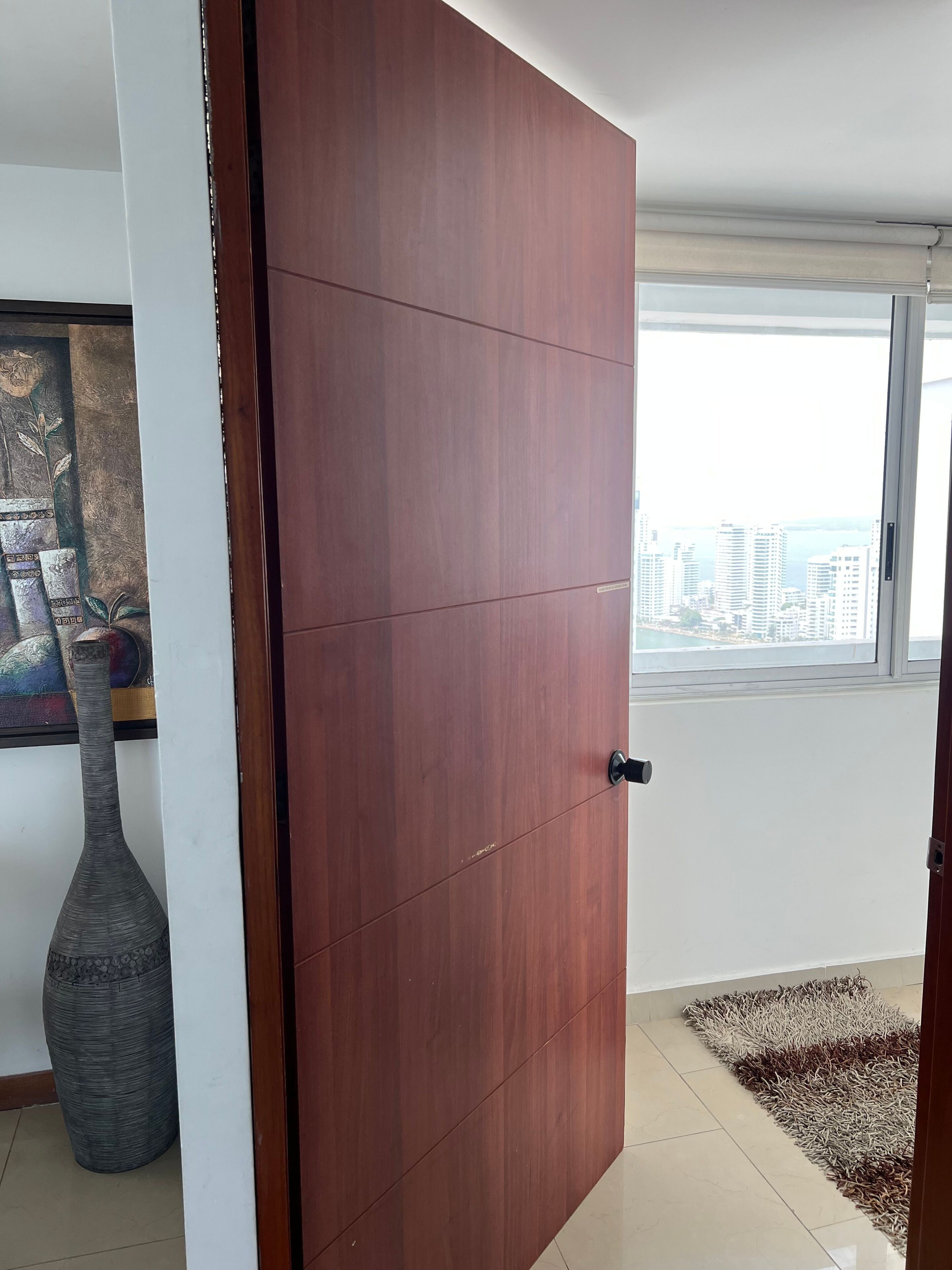 A room with a wooden door, a large window with a city view, a patterned rug, and a vase.
