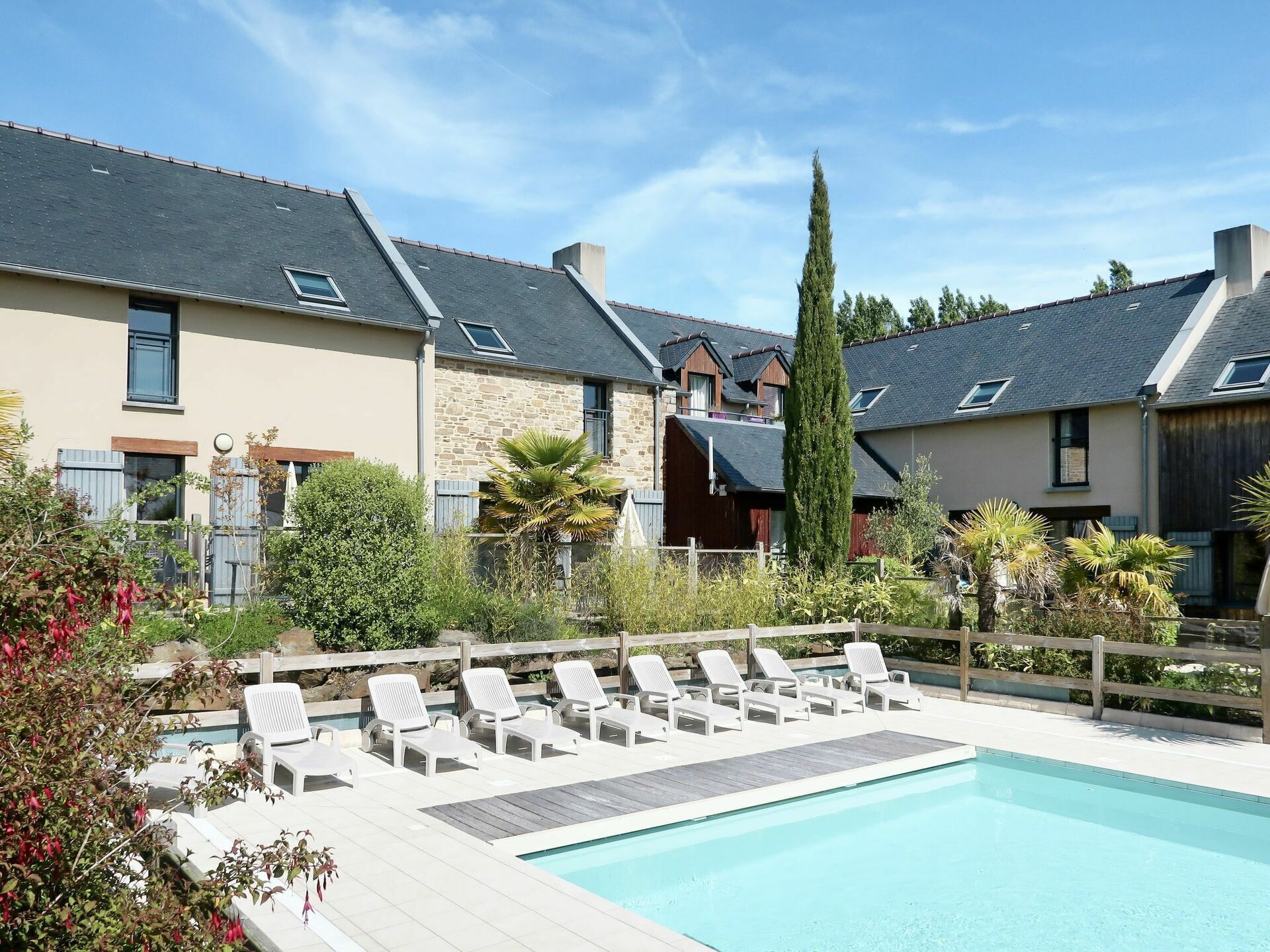 Une piscine entourée de chaises longues, elle-même bordée de maisons aux façades en pierre et aux toits en pente.