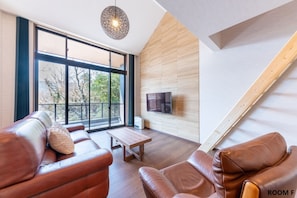 A modern living room with leather sofas, a wooden coffee table, a large window with a view of trees, and a mounted TV on the wall.