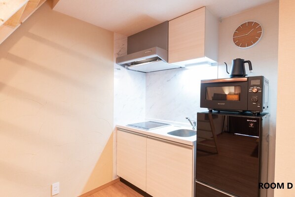 A compact kitchen with a microwave, sink, and cabinets. A clock is mounted on the wall.