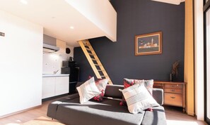 Un salon moderne avec un canapé, des coussins, une échelle en bois, une cuisine équipée et un tableau encadré au mur.