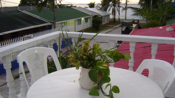 A round white table with two white chairs, a potted plant, and a view of a residential area with houses and a pink fence. - Room-green Sea Inn San Luis (San Andrés)
