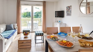 A modern living room with a sofa, a dining table set with seafood, and a TV mounted on the wall. - Beautiful apartment in villa for 4 guests with pool, hot tub, TV, terrace and pets allowed (Château-d'olonne)