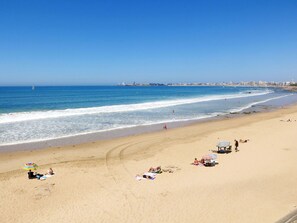 Ein Strand mit badenden und sonnenden Menschen, ein Segelboot in der Ferne und eine Stadtansicht im Hintergrund.